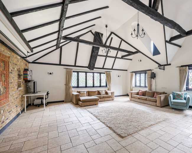 The image shows a spacious living area with high ceilings, exposed beams, large windows, and a mix of seating options, including sofas and armchairs, on a stone-tiled floor.