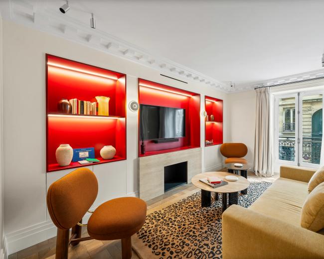 The image shows a modern living room with a beige sofa, orange accent chairs, a circular coffee table, and a wall-mounted television surrounded by red-lit shelves.