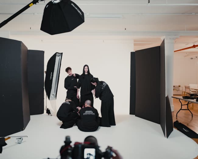A group of six individuals in black clothing is positioned in a studio setting, surrounded by large backdrops and lighting equipment.