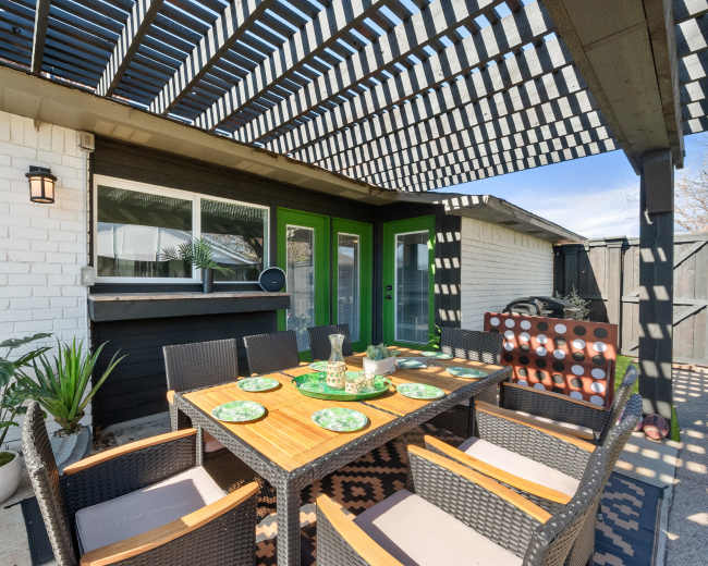 A patio area features a table set for dining, surrounded by chairs, beneath a slatted roof with green accents on the walls.