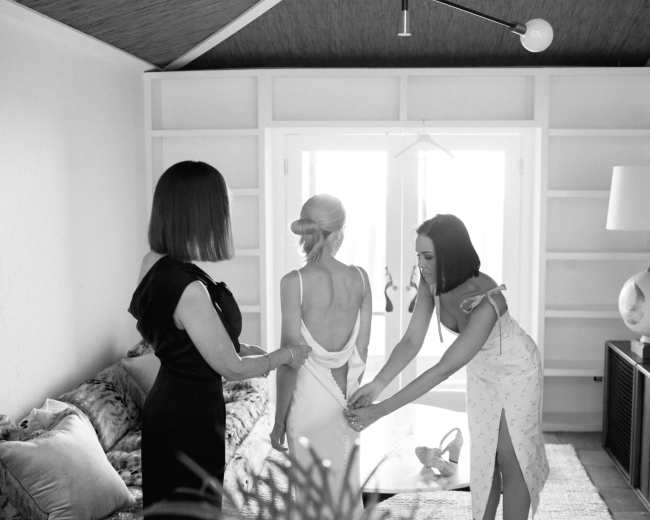 Three women are adjusting a wedding dress in a well-lit room with a sofa and large window.