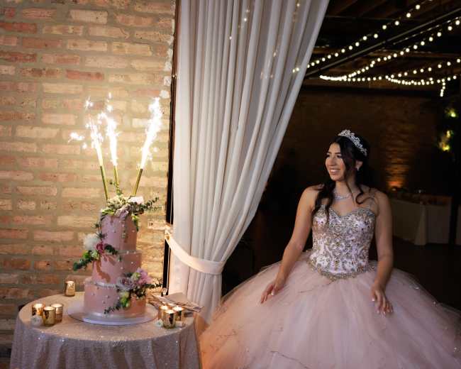 A woman in a formal gown stands next to a pink wedding cake adorned with sparklers in a decorated venue.