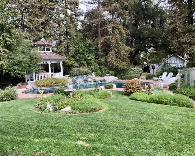 The image shows a landscaped backyard featuring a swimming pool, a gazebo, and several garden areas with greenery and flowers.