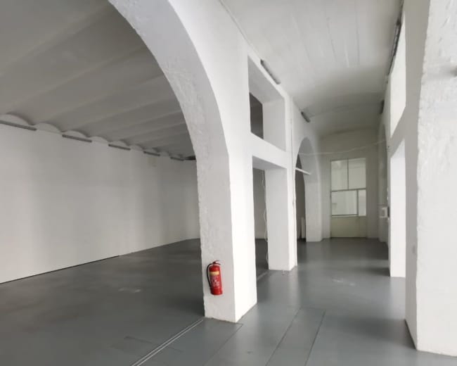 The image shows a spacious, vacant room with high ceilings, white walls, and a gray floor, featuring arched columns and a fire extinguisher on one wall.