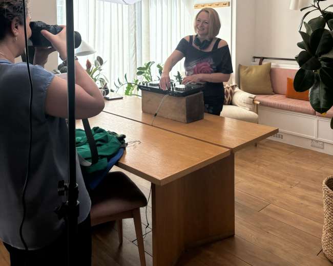 A woman stands behind a wooden table with a vintage item while another person takes her photo using a camera with a large softbox light.