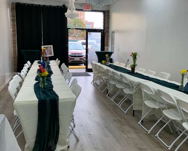 The image shows a decorated room with two long tables lined with white chairs, centered around a dark green table runner and flower vases, and illuminated by chandeliers.