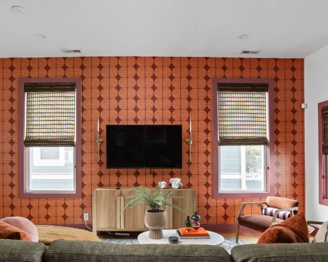 The image shows a living room with a patterned orange wallpaper, two windows with woven shades, a flat-screen TV mounted on the wall, and a sofa with cushions.