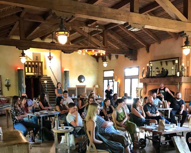 A group of people sits in a rustic room with wooden beams, engaged in a presentation or discussion.