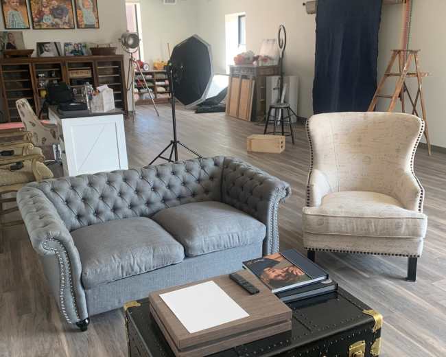 The image shows a modern studio space featuring a gray tufted sofa, a patterned armchair, a black trunk table, and various photo studio equipment.