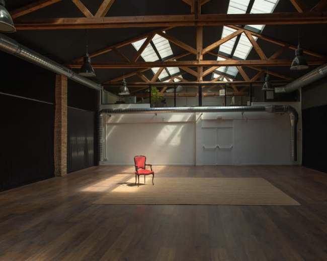 The image shows a spacious, empty room with wooden beams, a single red chair on a rug, and large windows allowing natural light to enter.