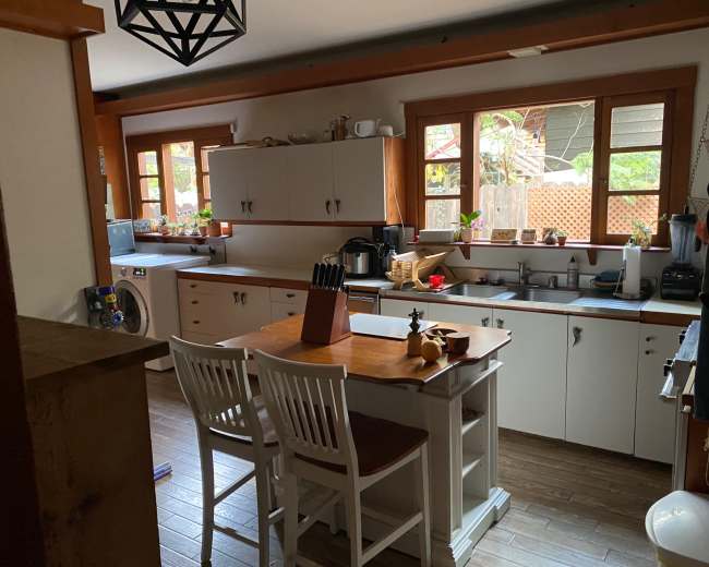 The image shows a kitchen with white cabinets, a wooden table with chairs, and windows allowing natural light to enter.