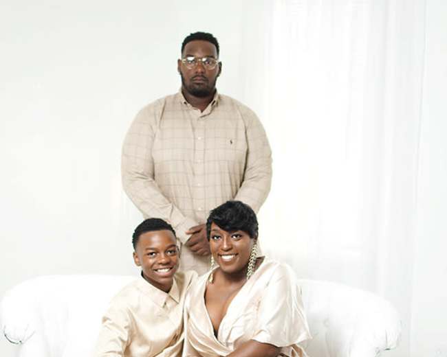 A man and woman, both dressed in formal attire, are seated on a white couch, while another man stands behind them.