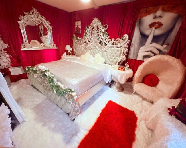 The image shows a lavishly decorated bedroom featuring a large ornate bed, plush white rugs, and red accents throughout the space.