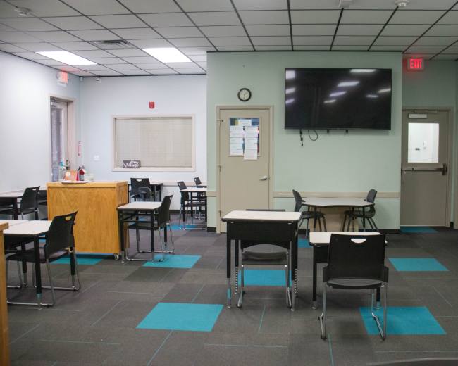 The image shows an empty classroom with desks arranged in rows and a large screen mounted on the wall.