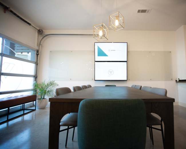 A conference room features a large table surrounded by chairs, with a wall-mounted screen displaying a presentation and decorative lighting above.