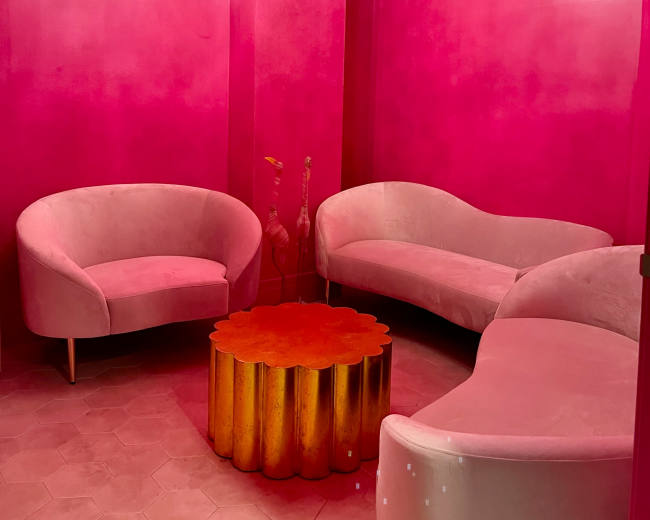 A room with bright pink walls, featuring two curved sofas and a round gold table in the center, illuminated by a modern chandelier.