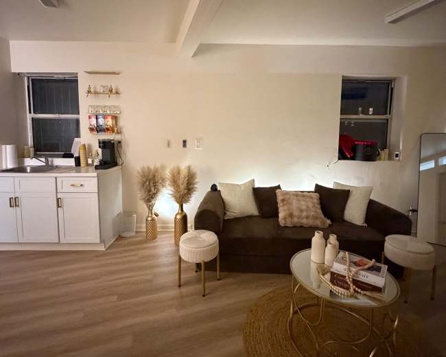 The image shows a cozy, softly lit living area featuring a dark sofa, two white side tables, a round rug, and a kitchenette in the background.