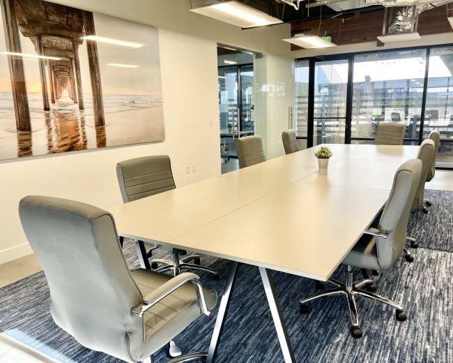 A modern conference room features a large, rectangular table surrounded by ergonomic chairs, with a seaside photograph on the wall and large windows overlooking an outdoor area.