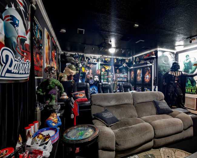 The room features a home theater setup with plush seating, walls adorned with superhero memorabilia, and various collectibles displayed throughout.
