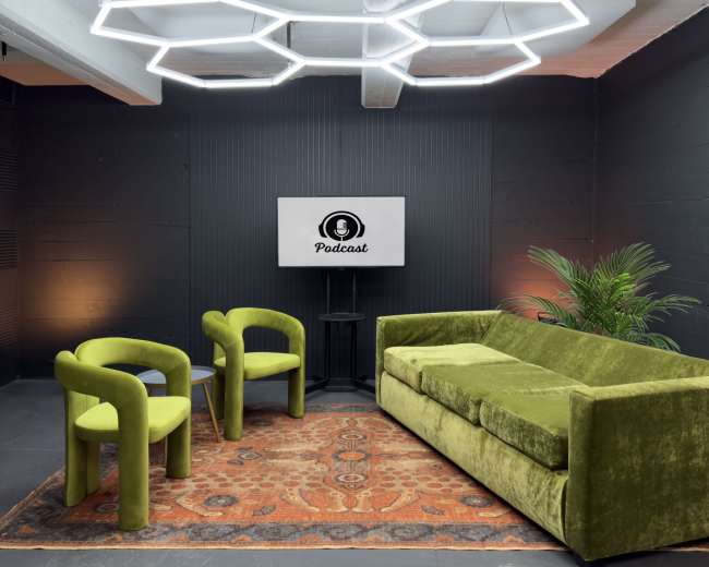 The image shows a modern podcast recording studio featuring a green velvet sofa, two matching chairs, a decorative rug, a potted plant, and a monitor displaying the word "Podcast."