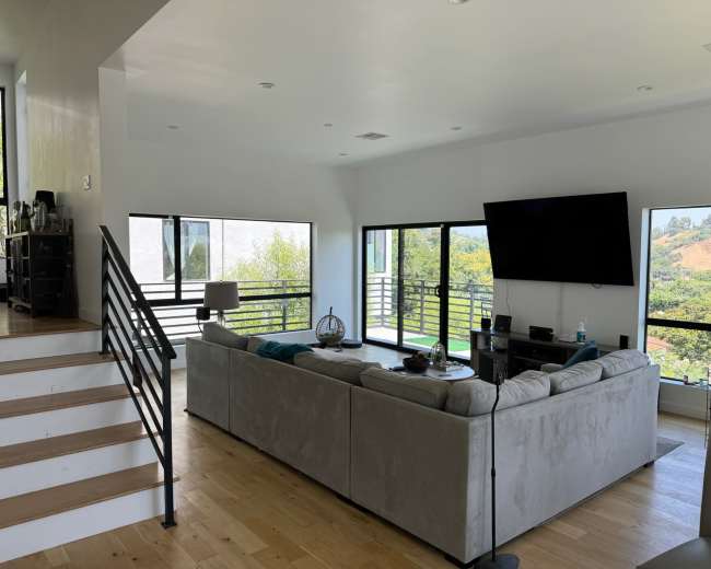 The image shows a modern living room with large windows, a sectional sofa, and a staircase leading to an upper level.
