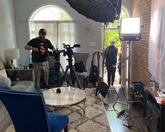 A film crew sets up equipment in a well-lit living room with large windows and a patterned rug.