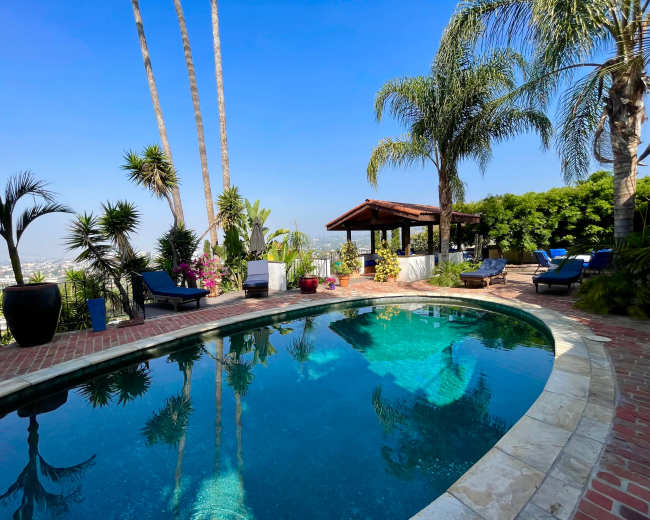 The image shows a luxurious swimming pool surrounded by palm trees and tropical plants, with a shaded patio area in the background.