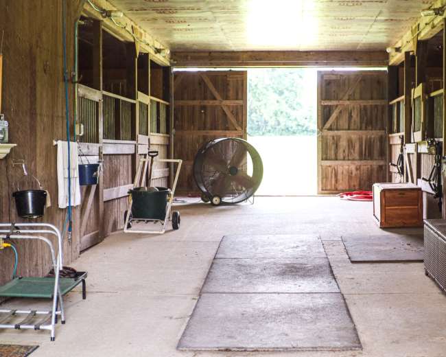 The image shows the interior of a barn, featuring several empty horse stalls, a large fan, and double doors leading outside.