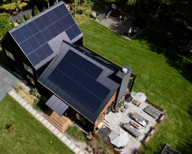 The image shows a modern house with a black exterior and several solar panels on its roof, surrounded by a landscaped yard with a patio and outdoor seating.