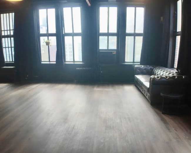 The image shows a spacious room with large windows, hardwood floors, and a black leather couch along one wall.