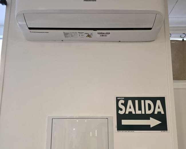 The image shows a wall-mounted air conditioning unit above a white electrical box and a telephone, with a green exit sign indicating "SALIDA" to the right.