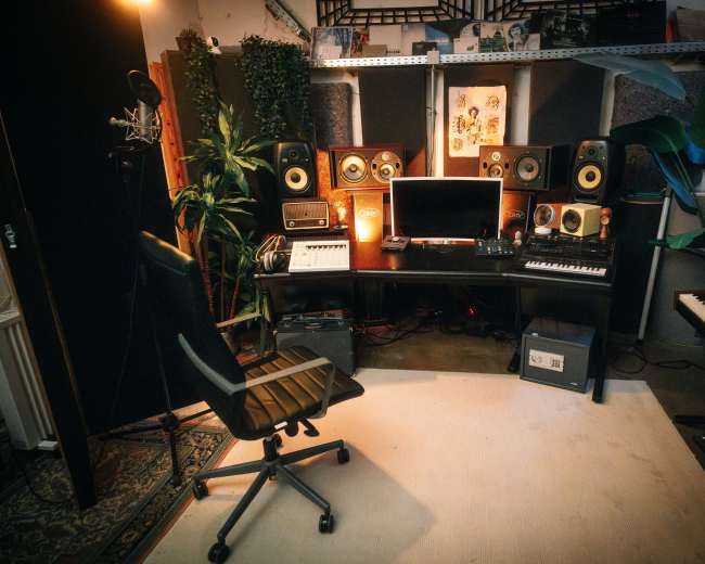 A music production studio features a desk with monitors, a keyboard, and various audio equipment, surrounded by plants and acoustic panels.