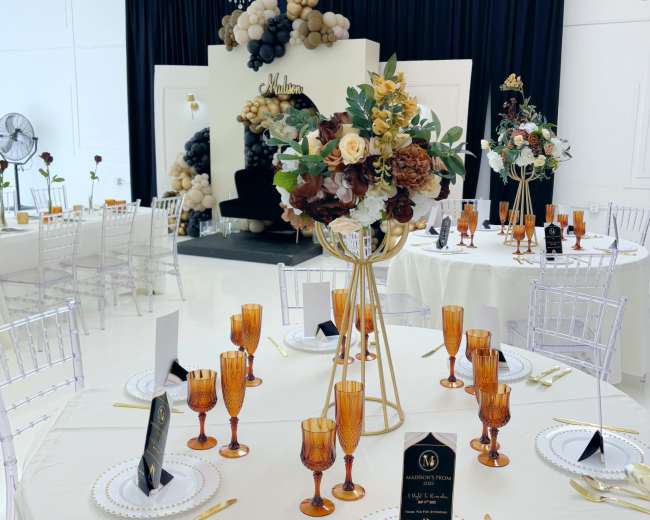 The image shows a banquet setup featuring round tables adorned with gold cutlery and amber glasses, surrounded by elegant decor including a floral centerpiece and a backdrop of balloons.