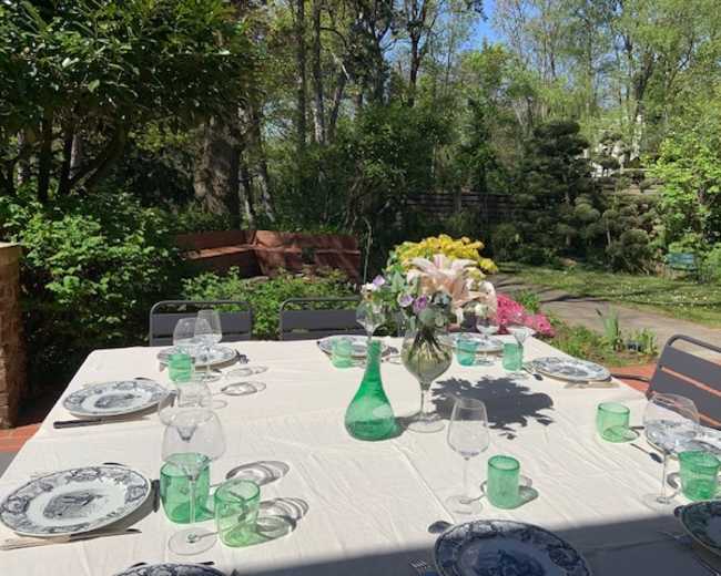 A dining table set with glassware and floral arrangements is positioned outdoors in a lush garden.