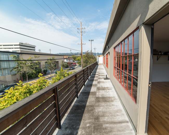 A narrow walkway with a wooden railing runs alongside a building, offering a view of neighboring structures and power lines against a clear sky.