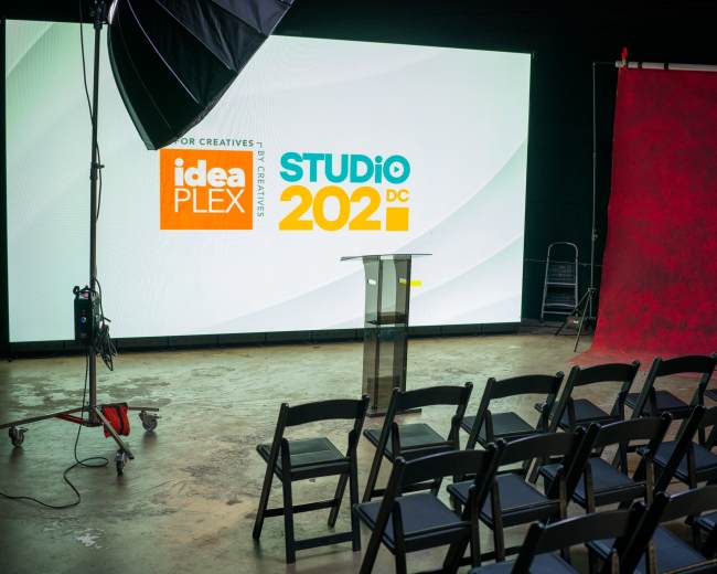 A stage is set with a podium in front of a large screen displaying "idea PLEX STUDIO DC 2021," with black folding chairs arranged in rows.