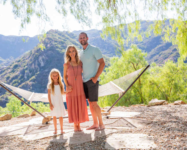 A family of three stands together in a scenic outdoor area with mountains in the background and a hammock nearby.