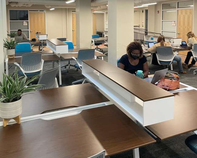 The image shows a modern, open workspace with several individuals seated at desks, some using laptops, and plants placed on the tables.