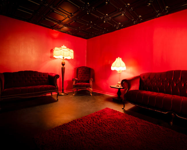 The image shows a room with red walls, two ornate sofas, a chair, and two lamps providing soft lighting.