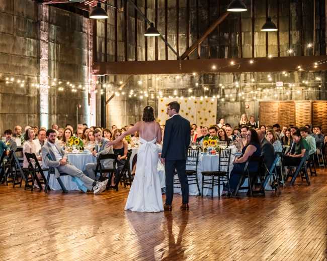 A couple stands together at the front of a rustic venue, addressing an audience seated at tables adorned with sunflower centerpieces.
