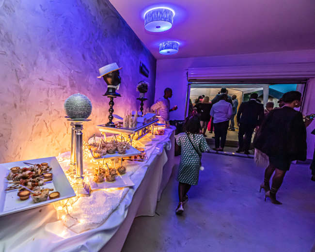 A buffet table adorned with various appetizers and decorative elements is set up in a dimly lit room, with guests mingling in the background.