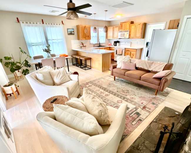The image shows a cozy living space with a kitchen and seating area, featuring light-colored furniture, wooden cabinets, and a fireplace.