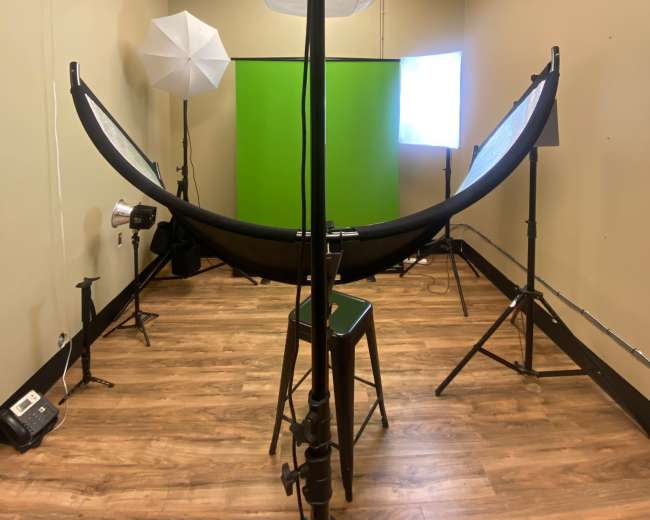 A studio setup featuring a green backdrop, various lighting equipment, and a stool in a room with wood flooring.