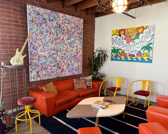 A vibrant living space features a colorful abstract painting, a large red couch, yellow chairs, and a coffee table on a black rug against a backdrop of exposed brick walls.