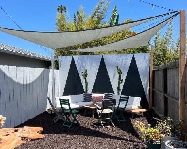 A shaded outdoor seating area with a geometric mural, surrounded by gravel and potted plants.