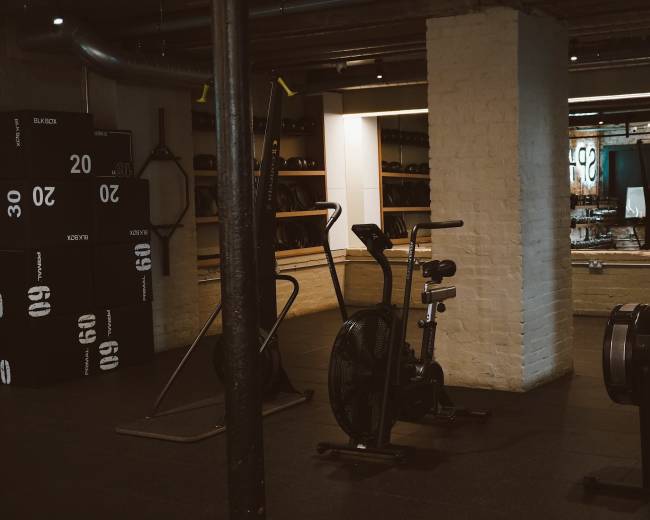 The image shows a fitness area with exercise equipment, including an air bike, rowing machine, and a few gym weights stacked on shelves.