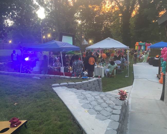 A gathering takes place outdoors with several tents, tables decorated with balloons, and a projector screen in the background.