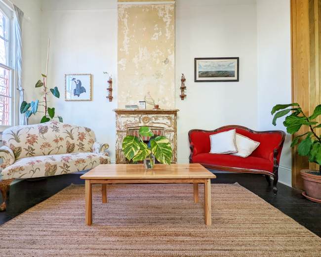 The scene features a cozy living room with a floral-patterned couch, a red vintage sofa, a wooden coffee table, and a plant by a decorative fireplace.