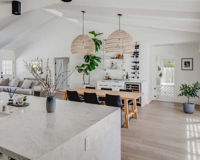 The image shows a modern open-concept living space featuring a kitchen with a marble countertop, a dining area with a wooden table, and a comfortable seating area.