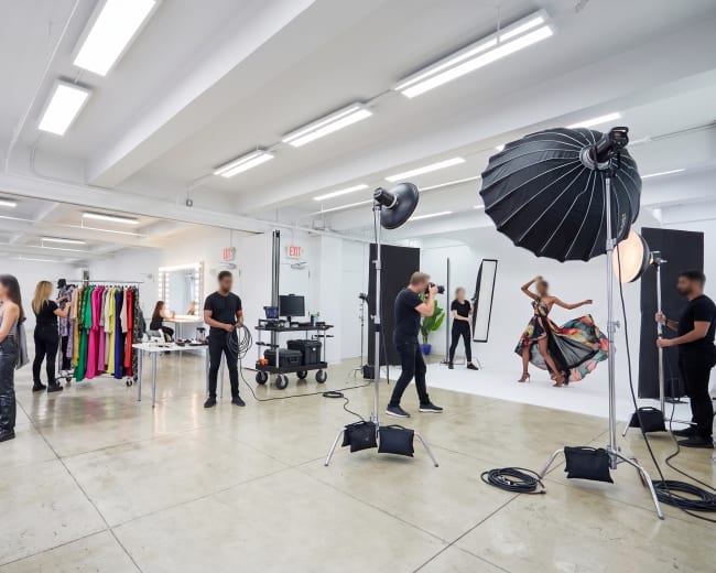 A photography studio features several people preparing for a photoshoot, with bright lighting, clothing racks, and a model posing against a white backdrop.